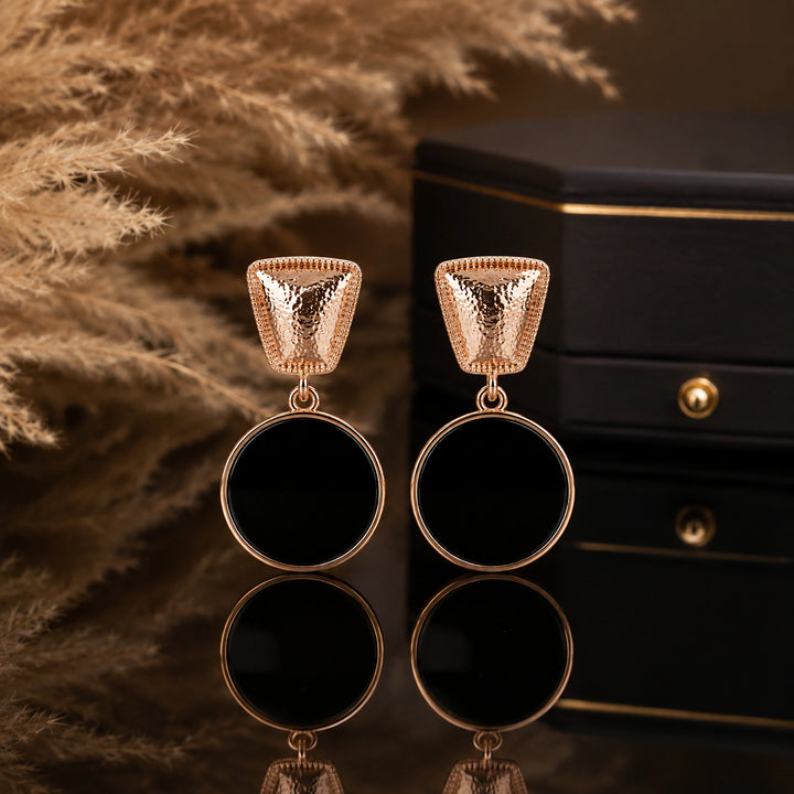 A pair of rose gold drop earrings with textured triangular top pieces and large circular black disc pendants, displayed on a reflective surface with a black jewelry box and dried pampas grass in the background.